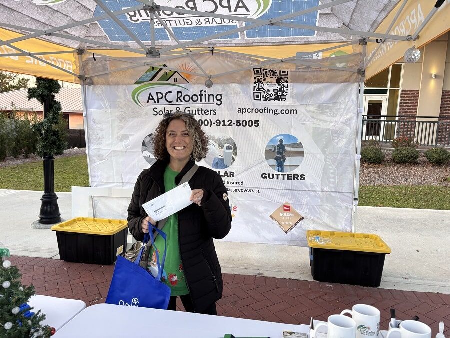 A woman stands in front of an APC Roofing booth, smiling and holding a white envelope and a blue bag at an outdoor event.
