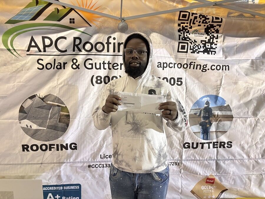 A man wearing glasses and a light hoodie stands in front of an APC Roofing banner, holding an envelope and facing the camera.