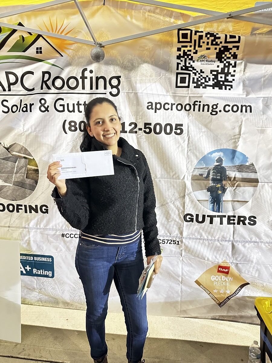 A woman stands in front of an APC Roofing banner, smiling and holding an envelope in her right hand.