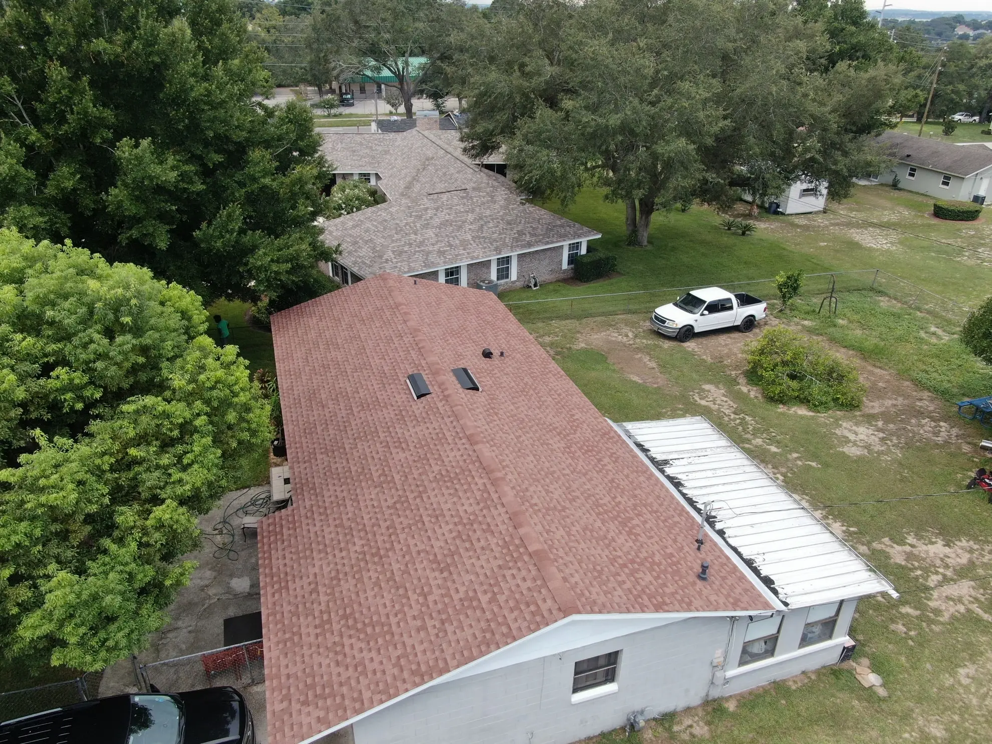 Aerial view of a house with a brown shingle roof, two skylights, a white truck parked nearby, trees, and other houses in the background. Aerial view of a house with a brown shingle roof, two skylights, a white truck parked nearby, trees, and other houses in the background.