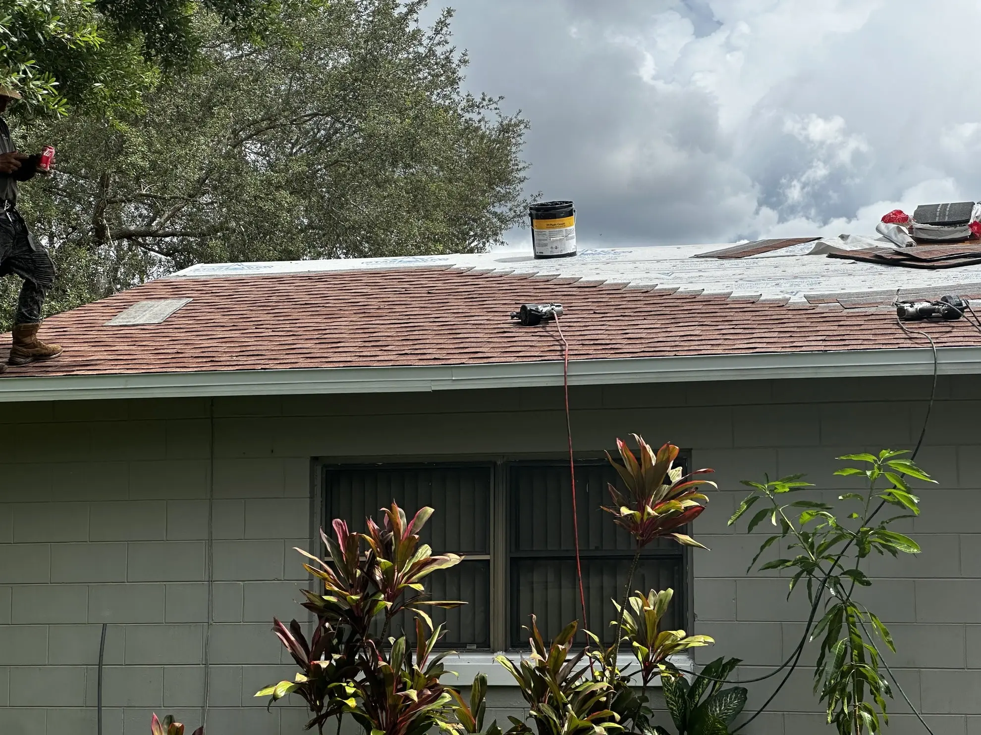 A person works on the roof of a house, with roofing materials and tools scattered on the shingles and some vegetation in the foreground. A person works on the roof of a house, with roofing materials and tools scattered on the shingles and some vegetation in the foreground.