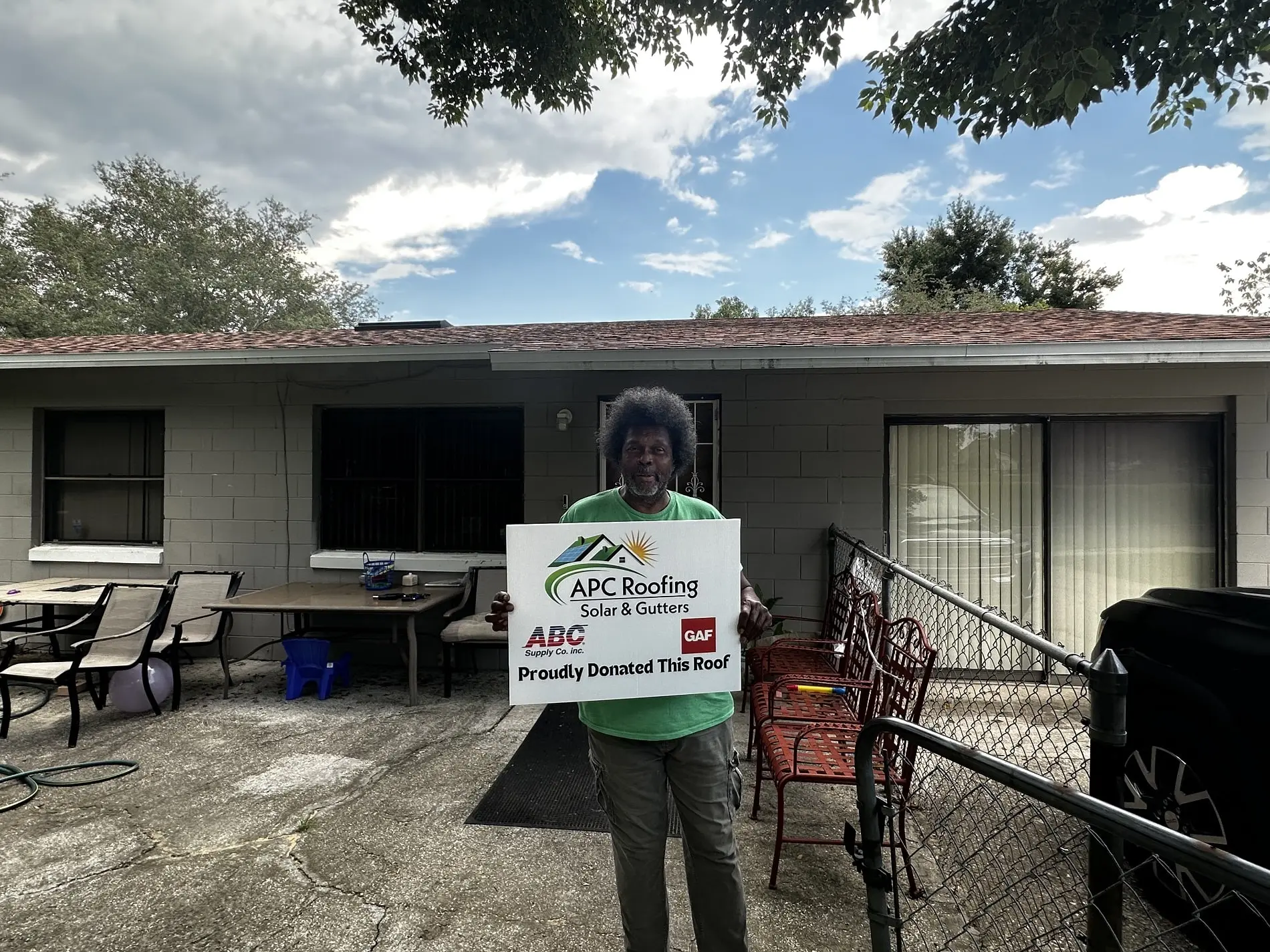 A person stands in front of a house holding a sign that reads, APC Roofing Solar & Gutters, Proudly Donated This Roof, with tables and chairs nearby. A person stands in front of a house holding a sign that reads, APC Roofing Solar & Gutters, Proudly Donated This Roof, with tables and chairs nearby.