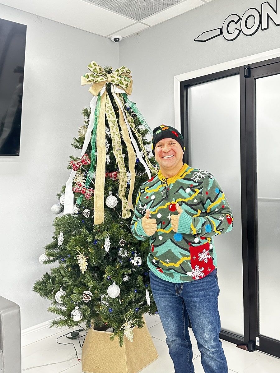 Man in a colorful holiday sweater and hat stands smiling with thumbs up next to a decorated Christmas tree indoors.