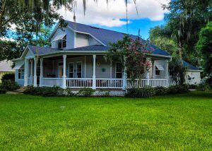 A white house with a large wraparound porch, surrounded by green grass, trees, and shrubs under a partly cloudy sky.