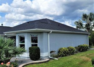A single-story house with light blue exterior walls, a dark shingle roof, white trim, and landscaped bushes, under a partly cloudy sky.