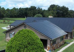 A large barn with a black metal roof sits on a grassy property surrounded by fenced pastures and trees, with horses visible in the background.