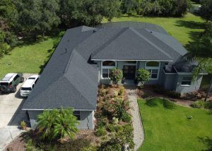 Aerial view of a large single-story house with a dark roof, two cars parked in the driveway, landscaped yard, and surrounding trees.
