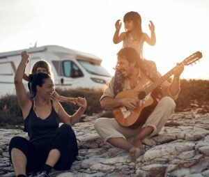 A man plays guitar while sitting on rocks, with a child on his shoulders. A woman and another child dance nearby. A camper van is parked in the background. A man plays guitar while sitting on rocks, with a child on his shoulders. A woman and another child dance nearby. A camper van is parked in the background.