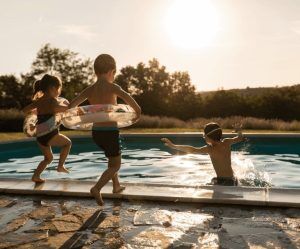 Three children with swim rings jump into an outdoor swimming pool at sunset, with trees and hills in the background. Three children with swim rings jump into an outdoor swimming pool at sunset, with trees and hills in the background.