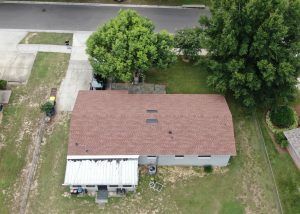 Aerial view of a single-story house with a brown roof, backyard, two large trees, and a partially covered patio. A driveway and street are visible at the top of the image.