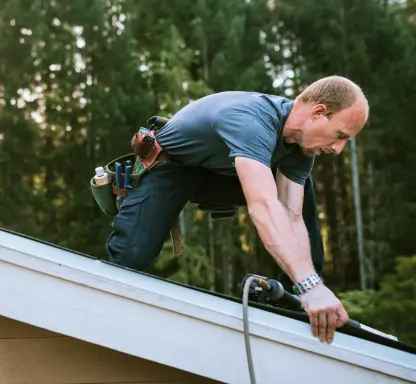 A man wearing a tool belt installs or repairs roofing materials on a sloped roof, using a tool while kneeling. Trees are visible in the background. A man wearing a tool belt installs or repairs roofing materials on a sloped roof, using a tool while kneeling. Trees are visible in the background.