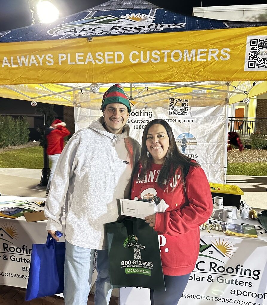 Two people stand smiling in front of an APC Roofing booth at an outdoor event, holding a gift card and a blue tote bag. The booth displays signs, cups, and promotional items.