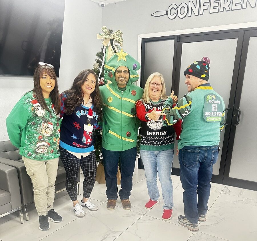 Five adults stand indoors, smiling and wearing festive holiday sweaters; one is dressed as a Christmas tree. A decorated tree and CONFERENCE sign are visible in the background.